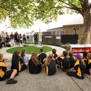 School children and community members gathered outdoors for the opening of the Garden of Memories, with illuminated artwork installed among the garden beds.