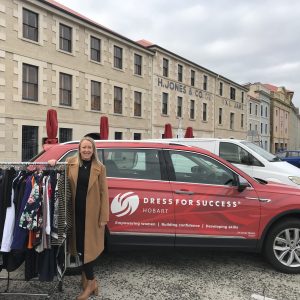Woman standing beside a rack of clothing and a Dress for Success Hobart vehicle outside a heritage building.