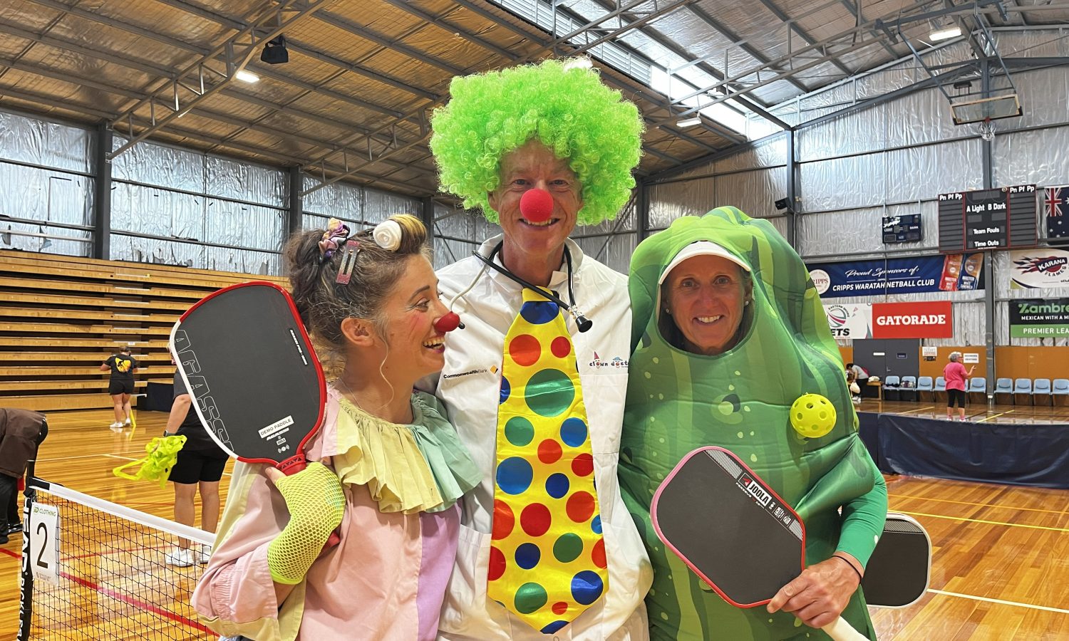 Three people in colourful costumes holding pickleball paddles and smiling together on an indoor court.