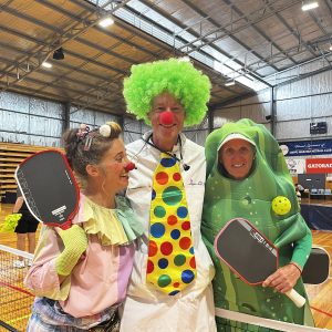 Three people in colourful costumes holding pickleball paddles and smiling together on an indoor court.