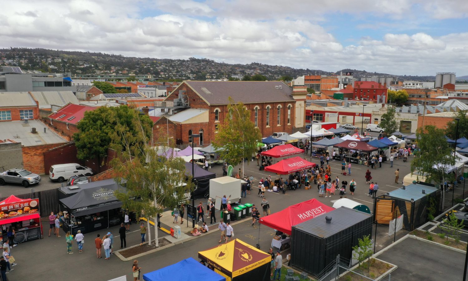 Elevated view across a busy Tasmanian community market, with people gathering among food stalls and colourful marquees surrounded by city buildings.