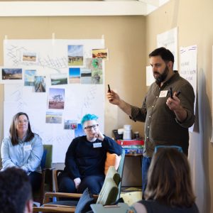 A facilitator speaking to a seated group during a Tasmanian Leaders retreat, with workshop notes, maps and photographs displayed around the room.